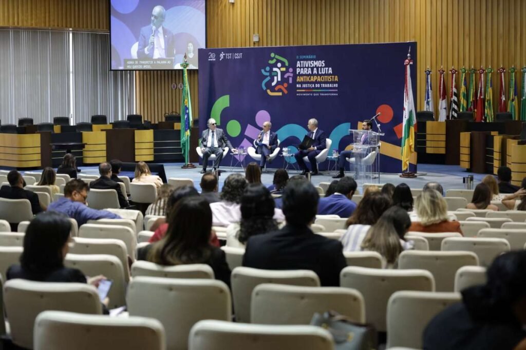 Foto da abertura do Seminário Ativismos para a Luta Anticapacitista. No palco, quatro homens estão sentados com banner do evento atrás.
