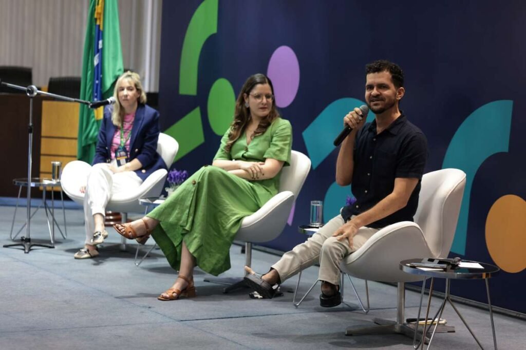 Foto de três participantes do seminário sentados em cadeiras lado a lado no palco.