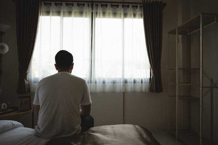 Homem sentado numa cama, olhando para janela fechada por cortina