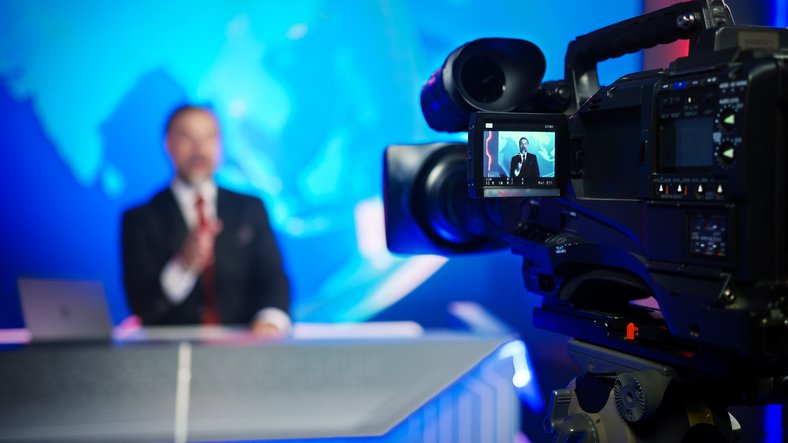 Apresentador em bancada de telejornal, diante de câmera de TV