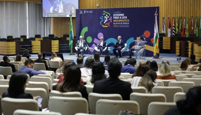 Foto da abertura do Seminário Ativismos para a Luta Anticapacitista. No palco, quatro homens estão sentados com banner do evento atrás.