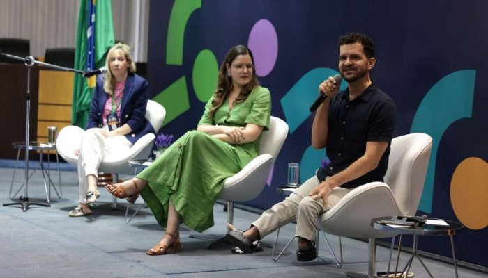 Foto de três participantes do seminário sentados em cadeiras lado a lado no palco.