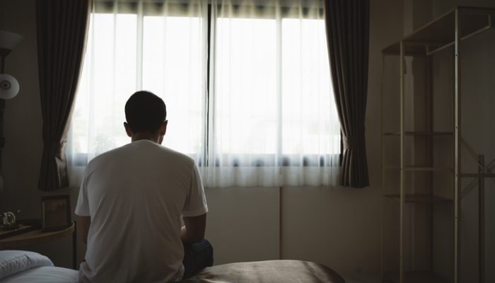 Homem sentado numa cama, olhando para janela fechada por cortina