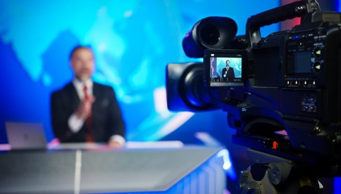 Apresentador em bancada de telejornal, diante de câmera de TV